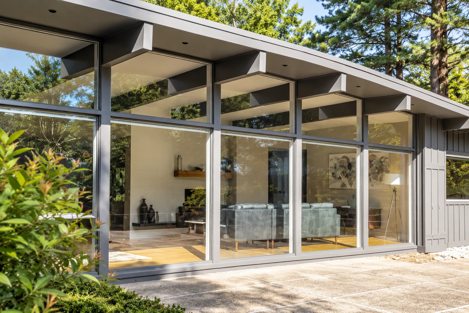 Modern four-season sunroom with floor-to-ceiling glass walls, gray framing, and a furnished living area among trees.