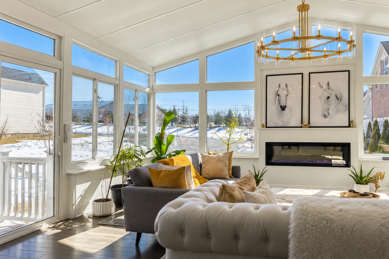 Bright all-season sunroom with tall windows overlooks snow-covered yards; a sofa, fireplace, and chandelier furnish the room.