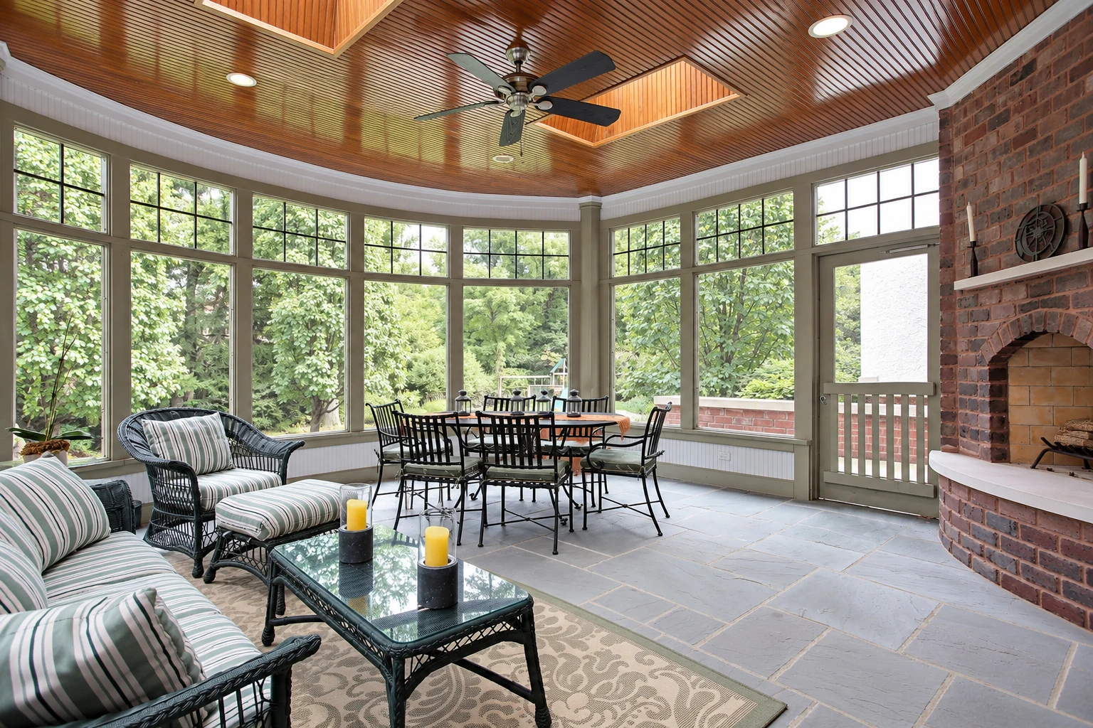 A custom sunroom with a wood-paneled ceiling, large windows, wicker seating, a dining table, and a brick fireplace.