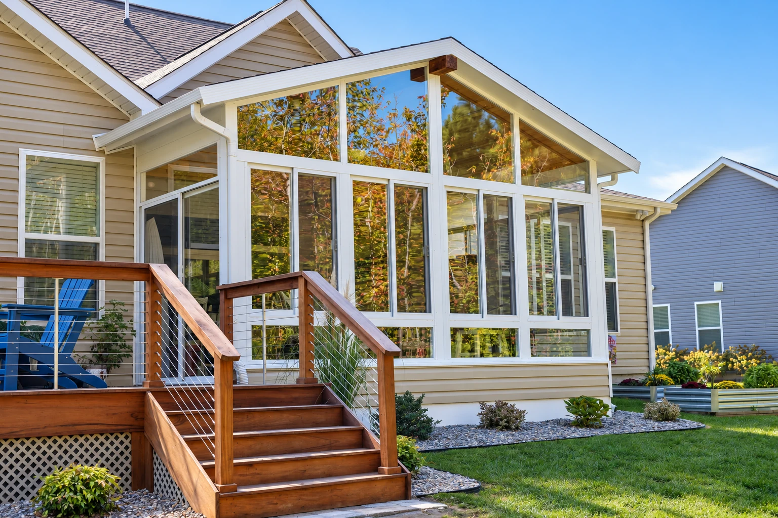Custom sunroom with a peaked glass gable and white frames adjoins a beige house above a wooden deck and stairs.