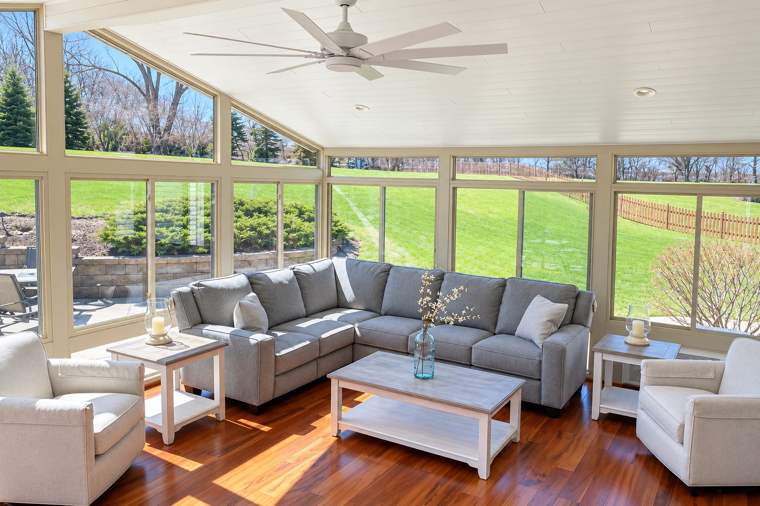 A bright sunroom with a gray sectional, white ceiling fan, and tall windows overlooking a green lawn.