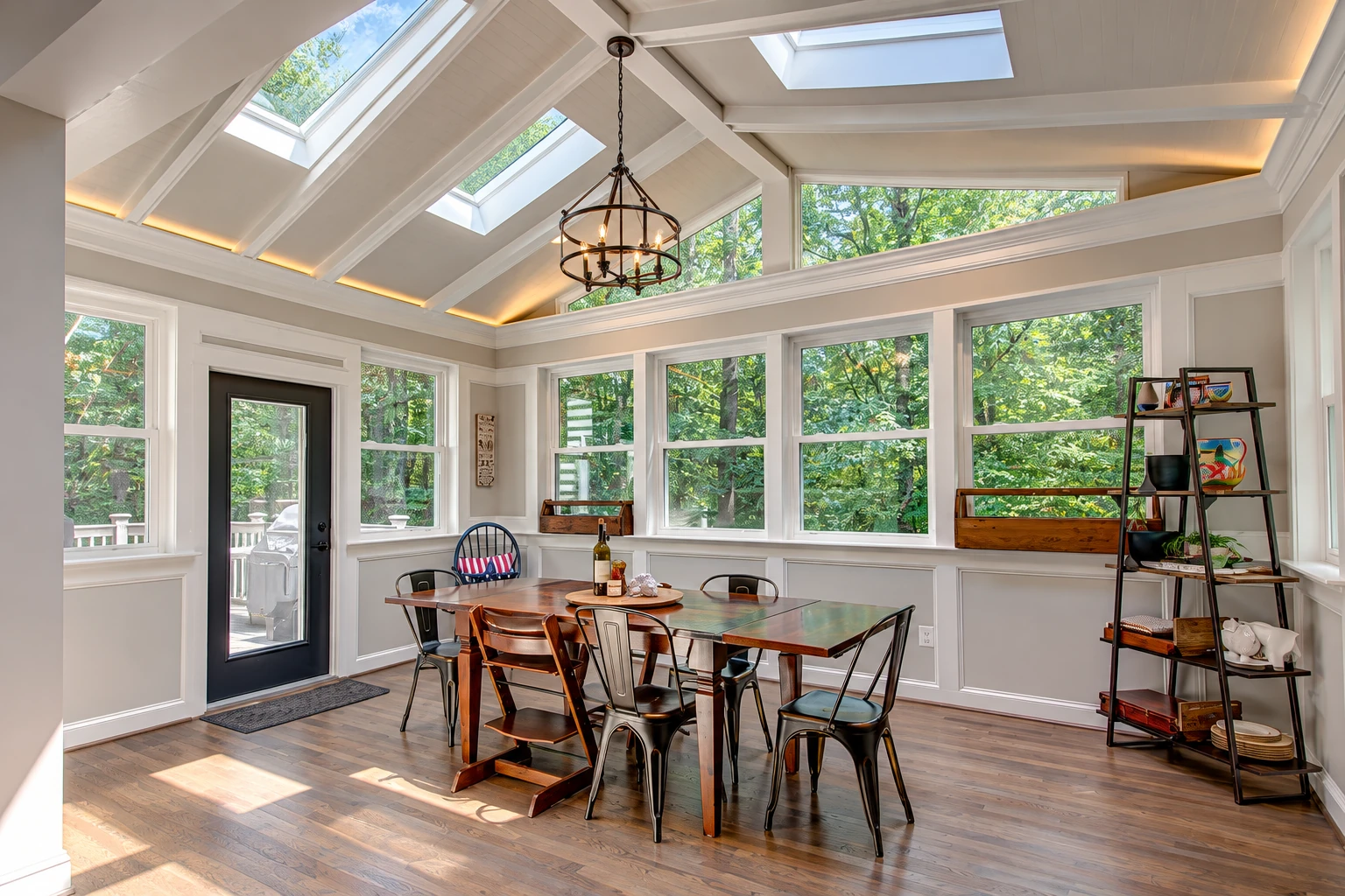 Sunroom dining area with broad windows and skylights features a wooden table, mixed chairs, and views of leafy trees.